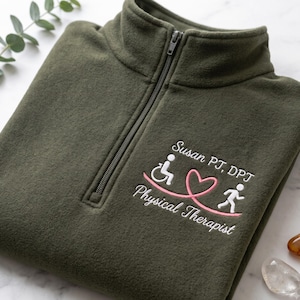 May include: An olive green quarter-zip fleece pullover featuring white and pink embroidery on the chest. The design includes the text "Susan PT, DPT" and "Physical Therapist" with a graphic of a wheelchair user and a runner connected by a heart.
