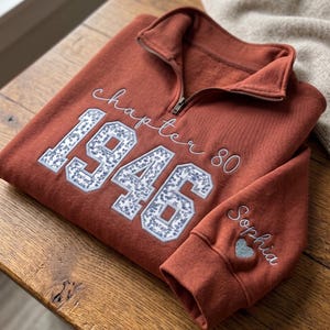 May include: A rust-colored quarter-zip sweatshirt featuring the embroidered text "chapter 80" and the large numbers "1946" filled with a blue and white floral pattern. The sleeve is embroidered with the name "Sophia" and a small blue heart.