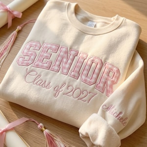 May include: A cream-coloured crewneck sweatshirt featuring the word SENIOR in pink and white gingham block letters, with the text Class of 2027 embroidered below in pink script and the name Michele embroidered on the sleeve cuff.