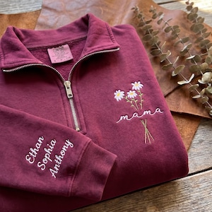 Peut inclure: Un sweat-shirt bordeaux à fermeture éclair quart, doté d'une fermeture éclair en métal argenté et d'une étiquette Comfort Colors au col. La poitrine présente un bouquet brodé de trois marguerites blanches avec le mot mama, et la manche porte les prénoms Ethan, Sophia et Anthony.