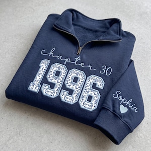 May include: A navy blue quarter-zip sweatshirt featuring embroidered text that reads "chapter 30" above the year "1996" in large block letters filled with a blue and white floral pattern. The sleeve is embroidered with the name "Sophia" and a heart.