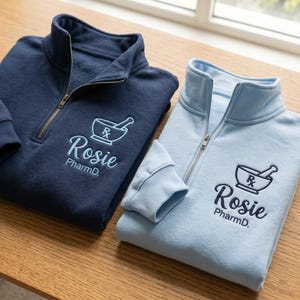 May include: A navy blue and a light blue quarter-zip sweatshirt, each featuring embroidered text that reads Rosie PharmD. below a mortar and pestle icon with an Rx symbol. Both garments have a high collar, a metal zipper, and ribbed cuffs.