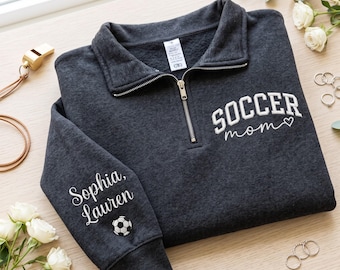 May include: A charcoal gray quarter-zip pullover featuring white embroidered text that reads "SOCCER mom" with a heart icon on the chest, and "Sophia, Lauren" with a soccer ball graphic on the sleeve. The garment has a fold-over collar and zipper.