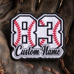 May include: A white embroidered patch featuring the number 83 in a block font with red baseball-style stitching details and black borders. Below the number, the text Custom Name is embroidered in a black cursive script on a white background.