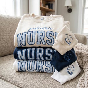 Peut inclure: Une pile de trois sweat-shirts à col rond en crème, bleu marine et blanc. Chacun porte le mot NURSE en lettres bloc à carreaux vichy bleus avec le mot pediatric brodé au-dessus. Les manches portent un stéthoscope en cœur et le nom Rosie.
