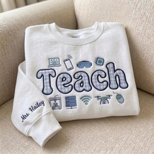 May include: A white crewneck sweatshirt featuring the embroidered word Teach in a blue and white gingham pattern. Surrounding the text are small embroidered technology icons including a laptop, phone, goggles, chip, printer, server, wifi, drone, and mouse. The sleeve is embroidered with Mrs. Hailey.