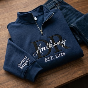 May include: A navy blue quarter-zip sweatshirt featuring embroidered text. The front displays the letters DR in black with the name Anthony in white script and EST. 2026 below. The sleeve is embroidered with the words General Surgery in white.