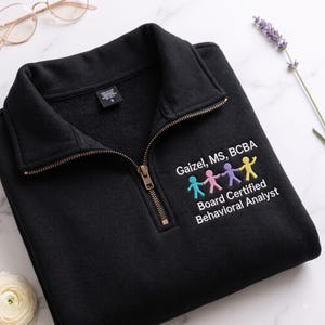 May include: A black quarter-zip sweatshirt featuring an embroidered design on the left chest. The text reads "Gaizel, MS, BCBA" and "Board Certified Behavioral Analyst" above a row of four small, colorful stick figures holding hands in teal, pink, purple, and yellow.