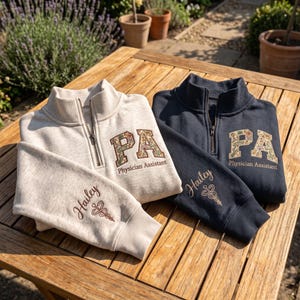 May include: Two quarter-zip sweatshirts, one light heather grey and one navy blue, featuring the embroidered letters PA with floral patterns and the text Physician Assistant. The sleeves are embroidered with the name Hailey and a caduceus symbol.
