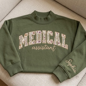 May include: An olive green mock neck sweatshirt featuring the word MEDICAL in large block letters filled with a floral pattern, the word assistant in cursive script, and the name Sophia embroidered on the sleeve in matching light thread.