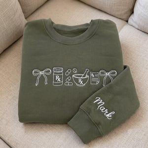 May include: An olive green crewneck sweatshirt featuring white embroidered line art of two bows, a prescription bottle, three pills, a mortar and pestle, and a prescription pad. The name Mark is embroidered in white script on the sleeve cuff.