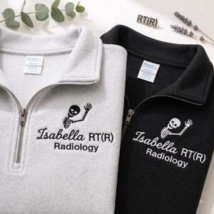 May include: A light gray and a black quarter-zip fleece pullover, each featuring an embroidered skeleton graphic and the text "Isabella RT(R) Radiology" on the chest. Both garments have a metal zipper, a collar, and a Port & Company brand label.