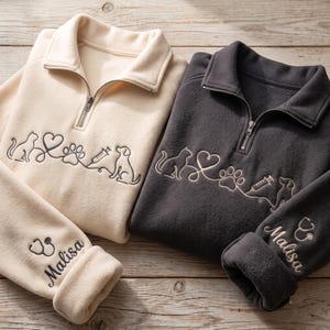 May include: A cream and a charcoal gray quarter-zip fleece pullover, each featuring an embroidered line art design of a cat, heart, paw print, syringe, and dog. The sleeve of each garment is embroidered with a stethoscope icon and the name Malisa.