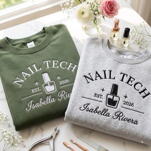 May include: An olive green sweatshirt and a heather gray sweatshirt, both featuring white and black embroidery respectively. The text reads NAIL TECH, EST. 2026, and Isabella Rivera, accompanied by a graphic of a nail polish bottle and stars.