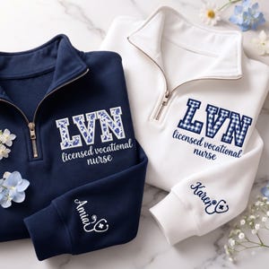 May include: A navy blue and a white quarter-zip sweatshirt. The navy sweatshirt features the text LVN and licensed vocational nurse with a floral pattern, and the name Amia with a stethoscope. The white sweatshirt features LVN in plaid.