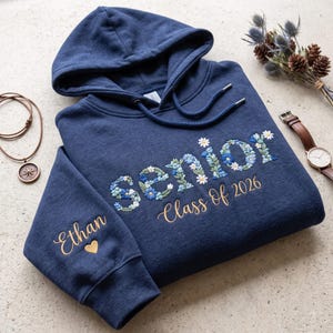 May include: A navy blue hooded sweatshirt featuring the word senior embroidered in a floral pattern with blue, white, and green thread. Below, the text Class Of 2026 is stitched in gold. The sleeve displays the name Ethan and a small gold heart.