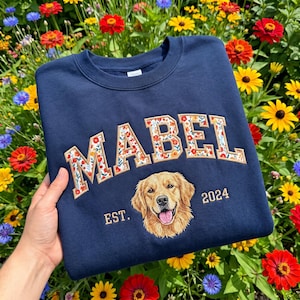May include: A navy blue crewneck sweatshirt featuring the name MABEL in large block letters filled with a colourful floral pattern. Below the name is an embroidered golden retriever portrait with the text EST. 2024 in gold thread.