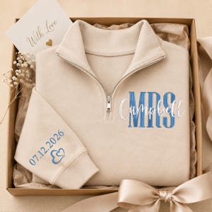 May include: A beige quarter-zip sweatshirt featuring blue embroidered text that reads MRS Campbell and the date 07.12.2026 with two small hearts on the sleeve. The garment is presented in a gift box with a card that says With Love.