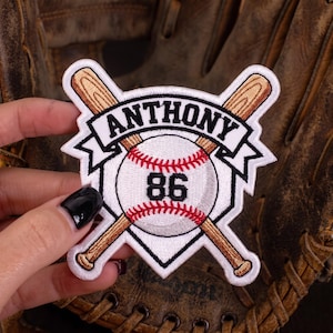 May include: A white embroidered patch featuring a baseball with red stitching and the number 86 in black. Two crossed wooden baseball bats are positioned behind the ball. A white banner across the top displays the name ANTHONY in black text.