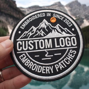 May include: A circular black embroidered patch featuring a white mountain range graphic, an orange sun, and the text EMBROIDERED IN SINCE 2023, CUSTOM LOGO, and EMBROIDERY PATCHES in white capital letters with a thick white stitched border.