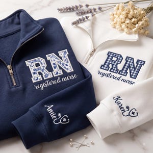 May include: A navy blue and a white quarter-zip sweatshirt, each featuring embroidered 'RN' and 'registered nurse' text. The navy sweatshirt has a floral pattern inside the letters and 'Amie' with a stethoscope on the sleeve. The white one has a blue gingham pattern and 'Karen' with a stethoscope on the sleeve.