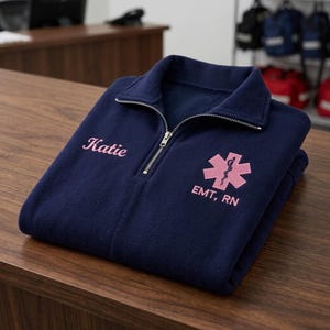 May include: A navy blue quarter-zip fleece pullover featuring light pink embroidery. The text Katie is embroidered on the right chest, and a pink Star of Life symbol with the text EMT, RN is embroidered on the left chest of the garment.