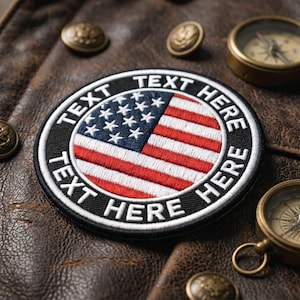 May include: A circular embroidered patch featuring the United States flag in the center with a black border. The white text "TEXT HERE" is stitched along the top and bottom edges of the black border, surrounding the red, white, and blue flag design.
