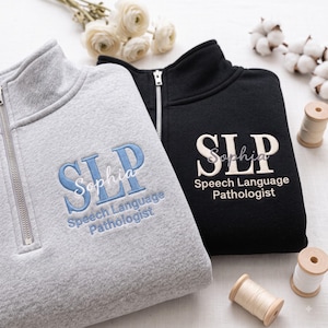 May include: A gray and a black quarter-zip sweatshirt, each featuring embroidered text that reads SLP, Sophia, and Speech Language Pathologist. The gray sweatshirt has blue embroidery, while the black sweatshirt has cream-colored embroidery.