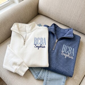 May include: A white and a heathered blue quarter-zip sweatshirt, each featuring a silver metal zipper. The white sweatshirt has light blue and dark blue embroidered text reading BCBA Sophia. The blue sweatshirt has white embroidered text reading BCBA Sophia.