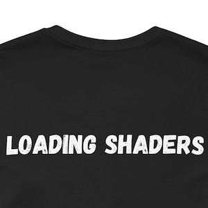 Puede incluir: Camiseta negra con el texto blanco "LOADING SHADERS" en una fuente en negrita y desgastada. El texto está centrado en la camiseta y el fondo es de color negro liso. La camiseta tiene cuello redondo.