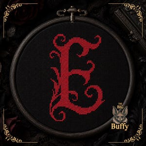 May include: A circular wooden embroidery hoop containing black fabric with a large, ornate, red cross-stitched letter E. In the bottom right corner, there is a small graphic of a bat with the text "THE BIG Buffy" printed in white lettering.