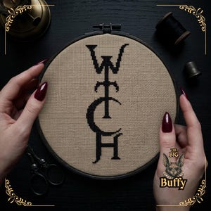May include: A circular embroidery hoop containing a cross-stitch design on beige fabric. The design features the word WITCH in black thread, with the letter C stylized as a crescent moon. The hoop is dark-colored and holds the fabric taut.