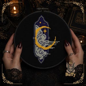 May include: A circular embroidery hoop containing a cross-stitch design on black fabric. The design features a yellow crescent moon and stylized grey clouds set within a vertical, dark blue, pointed frame with small yellow stars.