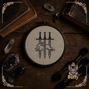 Könnte beinhalten: Ein runder Stickrahmen aus Holz mit einem Kreuzstichmotiv auf beigem Stoff. Das Design zeigt die schwarze Silhouette eines Hasen, der horizontal über drei vertikale schwarze Schwerter springt. Der Rahmen ist aus dunkelbraunem Holz.