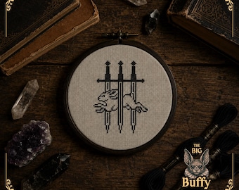 May include: A circular wooden embroidery hoop containing a cross-stitch design on light beige fabric. The design features a black silhouette of a rabbit leaping across three vertical swords. The text THE BIG Buffy is visible.