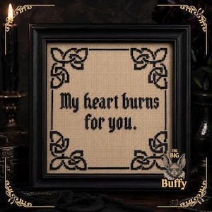 May include: A square cross-stitch embroidery in a black frame featuring the black text "My heart burns for you." centered on a beige fabric background. The design includes black geometric knotwork patterns in each of the four corners.
