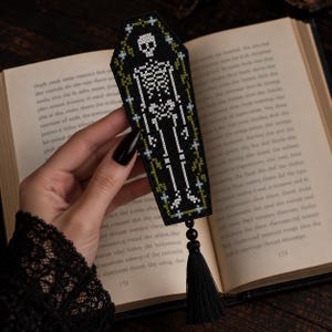 May include: A coffin-shaped bookmark featuring a white cross-stitched skeleton on a black background, framed by green vines and small blue flowers. The bookmark is finished with a black beaded accent and a long black tassel at the bottom.