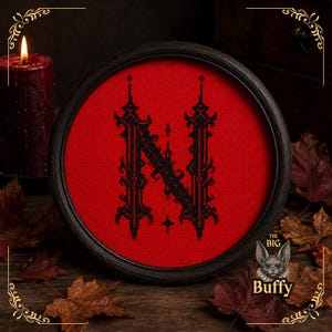 May include: A circular black wooden frame containing a cross-stitch embroidery of the letter N in black thread on a vibrant red fabric background. The letter features an ornate, gothic-inspired typeface with sharp, decorative edges and small crosses.