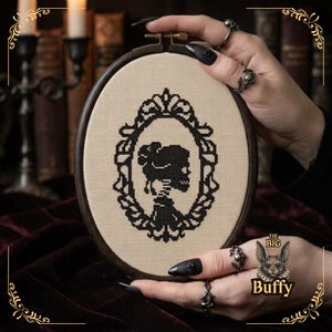 Op de afbeelding: Een ovale borduurring met een kruissteekontwerp op beige stof. Het ontwerp toont een zwart silhouet van het hoofd en de schouders van een persoon binnen een sierlijk, decoratief ovaal frame. De ring heeft een donkere houten afwerking en een metalen schroef.