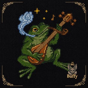 May include: A cross-stitch embroidery on black fabric featuring a green frog playing a brown lute. The frog has a blue smoke cloud above its head, yellow stars, and a musical note. The text "THE BIG Buffy" appears near a small bat illustration.