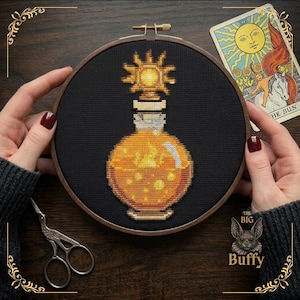 May include: A circular wooden embroidery hoop containing a cross-stitch design of a round glass bottle filled with glowing orange liquid. The bottle is topped with a golden sun emblem. The background fabric is solid black.