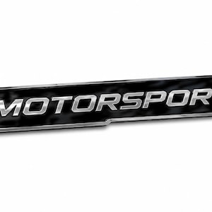 Op de afbeelding: Een rechthoekig embleem met een zwarte achtergrond en een metallic zilveren rand. Het woord MOTORSPORT staat in dikgedrukte, hoofdletter, metallic zilveren letters in het midden van het embleem. Het ontwerp heeft een slanke, schuine vorm.