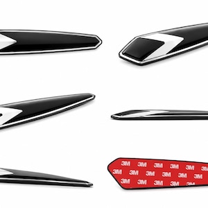 Könnte beinhalten: Ein Set aus länglichen, spitz zulaufenden Auto-Emblemen mit glänzend schwarzer Oberfläche und silbernem Chevron-Akzent. Die Embleme haben einen metallisch silbernen Rand und eine rote Kleberückseite mit dem wiederholten Text 3M.
