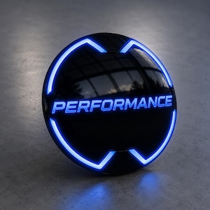 Pode incluir: Um emblema circular preto com a palavra PERFORMANCE em letras maiúsculas azul brilhante. O emblema é emoldurado por um anel de LED azul vivo que é interrompido por quatro entalhes distintos nas partes superior, inferior, esquerda e direita.