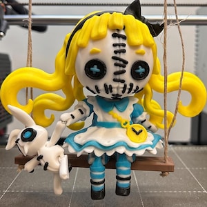 May include: A stylized doll with bright yellow hair, a black bow, and a white face featuring black stitched details and blue button eyes. The doll wears a blue and white dress with a yellow pocket watch accessory and holds a small white rabbit figure.