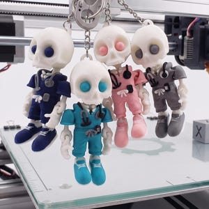 May include: A set of four articulated skeleton keychains, each wearing scrubs in different colors including navy blue, teal, pink, and gray. Each skeleton has a matching stethoscope around its neck and sneakers on its feet, all attached to a metal ring.