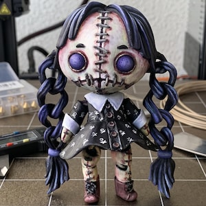 May include: A small, stylized figurine with a pale, stitched face, large purple eyes, and dark blue braided hair. It wears a black dress with a white collar and cuffs, featuring a white pattern, and dark boots. The figure has visible stitches.