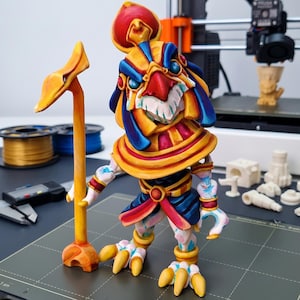 May include: A 3D printed figurine of an anthropomorphic bird deity wearing a golden headdress with red and blue accents. The figure holds a tall yellow staff and features a painted finish with vibrant yellow, red, and blue colors throughout.