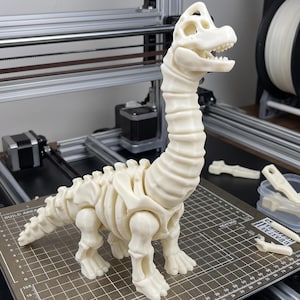 May include: An off-white, 3D-printed articulated dinosaur skeleton figure with a long neck, a detailed skull, and segmented body parts. The figure stands on a dark build plate with a grid pattern, accompanied by a few loose, matching plastic pieces.