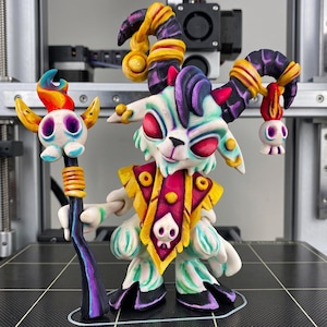 Op de afbeelding: Een 3D-geprint figuurtje van een gehoornd wezen met witte vacht, rode ogen en paarse accenten. Het figuur draagt een geel en magenta borststuk met een schedel-embleem en houdt een staf vast die is bekroond met een brandende schedel en bedel.